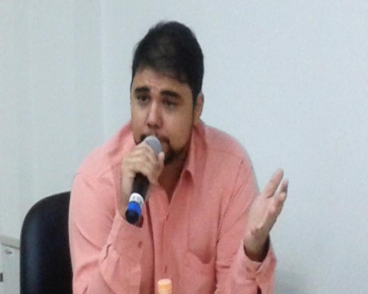 Homem de cabelos pretos curtos e barba gesticula falando ao microfone