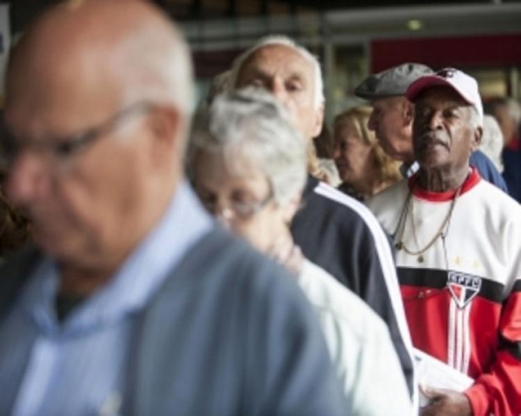 Fila com quatro idosos em primeiro plano, tendo na frente homem grisalho de óculos