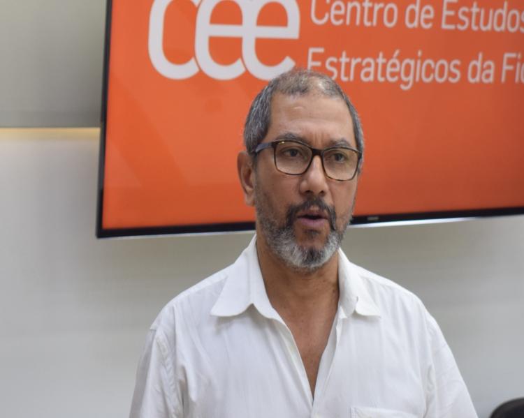 Homem de óculos e camisa branca, em primeiro plano, olha para frente e parece estar falando, tendo ao fundo tela de TV em cor laranja