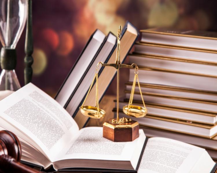 Livro aberto com uma pequena balança dourada sobre ele e uma pilha de livros ao fundo