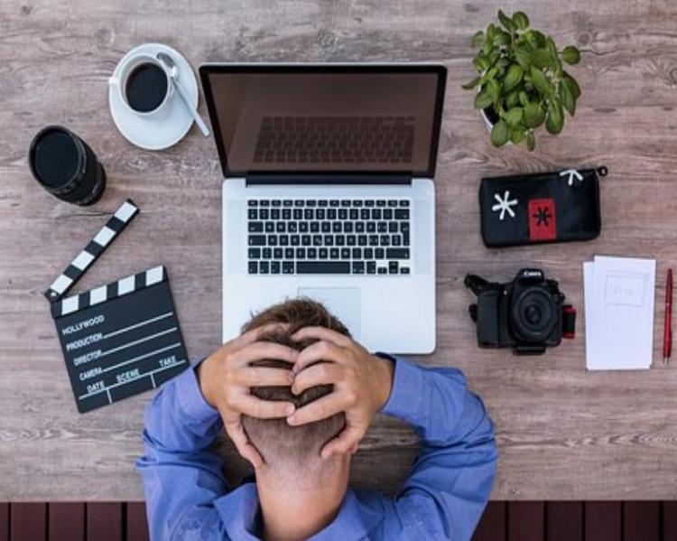 Homem de cabeça baixa com as mãos spbre a cabeça sentado a uma mesa com notebook, celular e outros equipamentos eletrônicos