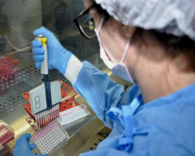 Mulher em um laboratório, de perfil, com touca e máscara de proteção, manipula um tubo de ensaio