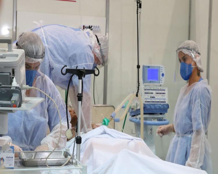 Profissional de saúde com equipamentos de proteção observa paciente monitorizado em leito hospitalar