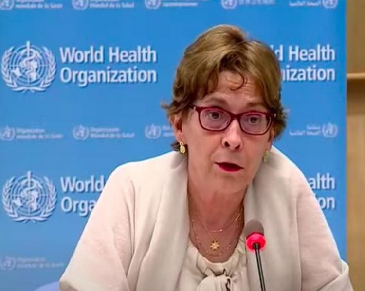 Mjulher de cabelos claros curtos e óculos, usando roupa clara, fala em uma mesa de debates, tendo como fundo um painel azul com a logomarca World Health Organization