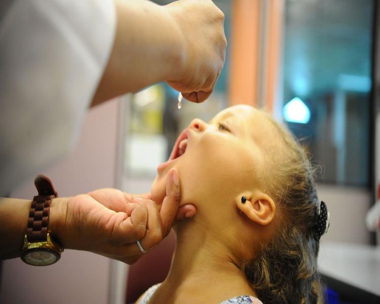 Rosto de menina com a boca aberta, recebendo uma gota de vacina de mãos femininas