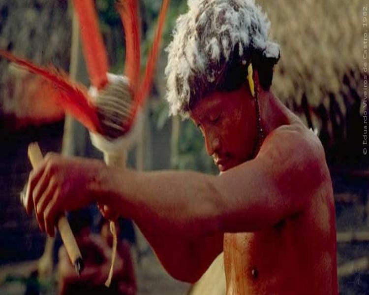 Homem indígena, de lado, olhando para baixo, segurando um instrumento de ritual xamânico
