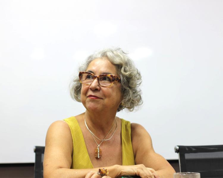 Mulher de cabelos grisalhos, óculos e blusa amarela, tem os braços apoiados sobre a mesa e olha o lado