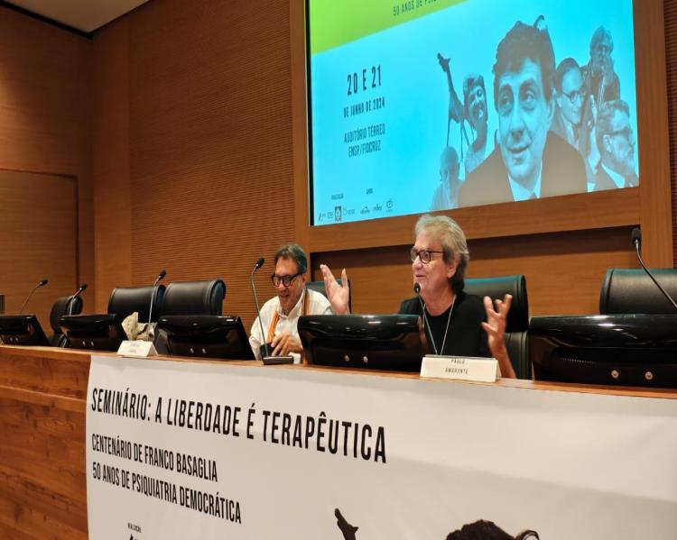 Dois homens em uma mesa de debates, um deles fala ao microfone. No telão, atrás, imagem de cartaz do seminário, com foto de Franco Basaglia