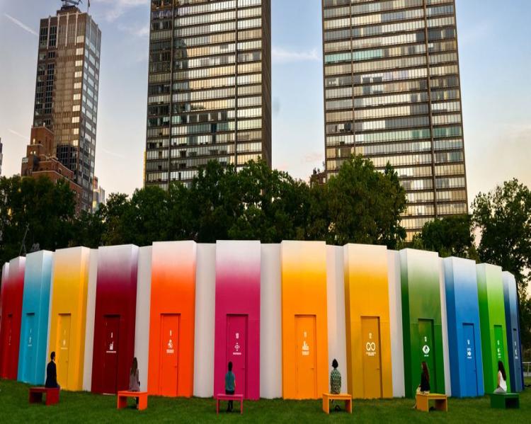 Um círculo de portas coloridas, observadas por pessoas sentadas em bancos, circunda três prédios altos
