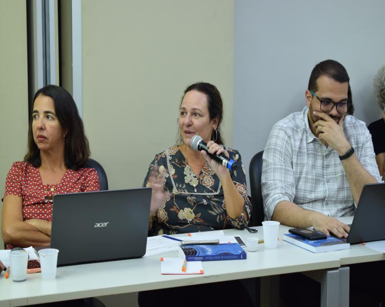 Duas mulheres e um homem estão em uma mesa de debates, ele fala ao microfone