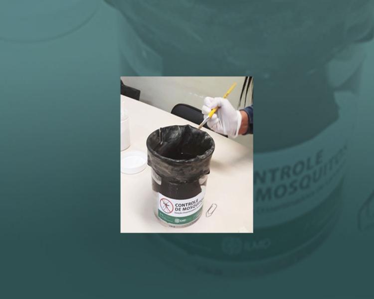 Mão com luva, segurando um pincel, está próxima à borda de um recipiente de cor preta, com rótulo onde lê Controle de Mosquitos, com o desenho de um mosquito ao lado